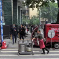 Woman pushing a cart loaded with large pots along the roadside while pedestrians and traffic pass by