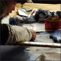 An embroiderer leans closely over a frame while working with fine dark silk threads.
