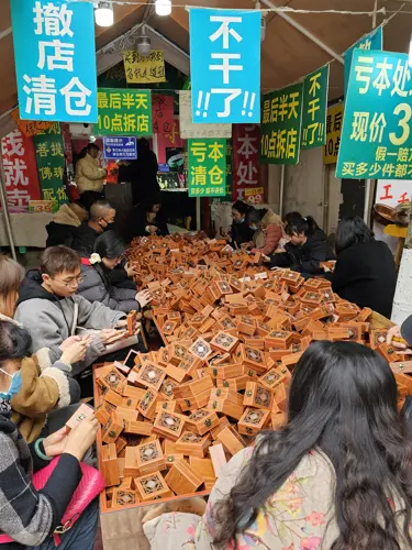 Street market stall covered with hundreds of small wooden “blind boxes,” as shoppers sit around opening them to search for small inexpensive items like jewelry and keychains.