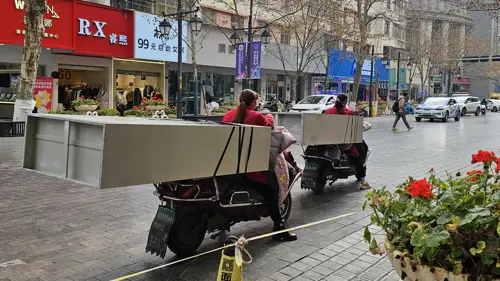 Two women riding motorbikes while transporting tall furniture pieces strapped to the back of their bikes.