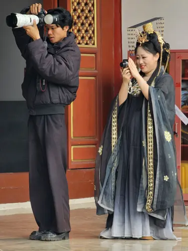 Male photographer with large professional camera and a woman in an elaborate ancient-style costume photographing a model out of frame.
