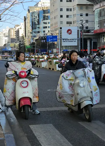 Two women riding electric scooters in a Chinese city, both using large quilted scooter blankets wrapped around the handlebars and their legs to stay warm.