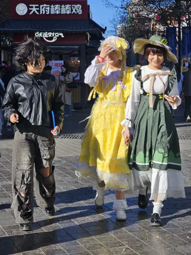 Three young people walking on a sunny Chinese street, two wearing frilly Lolita-style dresses and one in dark alternative streetwear influenced by anime and cosplay culture.