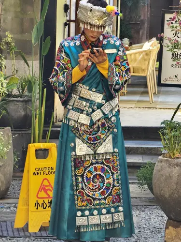 Woman in an elaborate ethnic minority costume looking at her mobile phone near a yellow wet-floor sign.