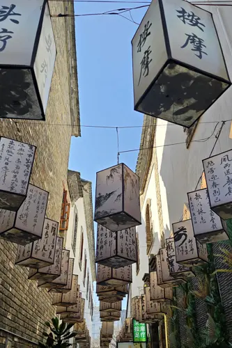 Narrow pedestrian lane in Kunming decorated with cube-shaped lanterns painted with Chinese calligraphy, hanging between old brick and plaster buildings under a blue sky.