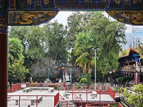 Stepped rectangular pond at Wenmiao in Kunming, enclosed by red metal railings and stone benches, viewed through the painted wooden beams of the temple pavilion.
